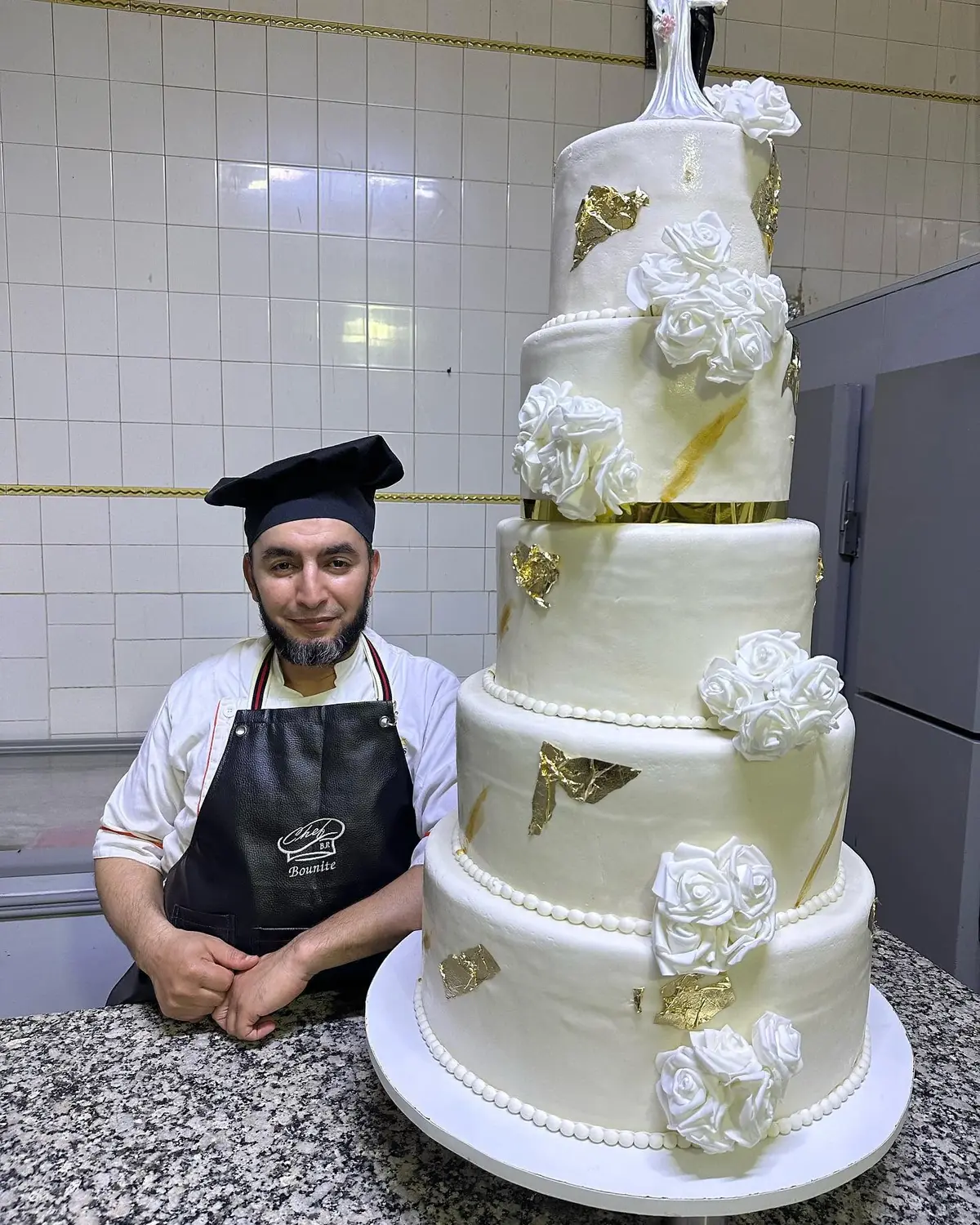 Le chef Bounite dans son atelier carrelé, en tablier brodé, à côté d'une pièce montée ivoire de cinq étages ornée de roses en sucre et de papillons dorés.
